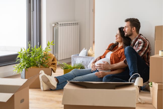 A couple sitting on the floor both holding onto the pregnant belly surrounded by cardboard boxes 